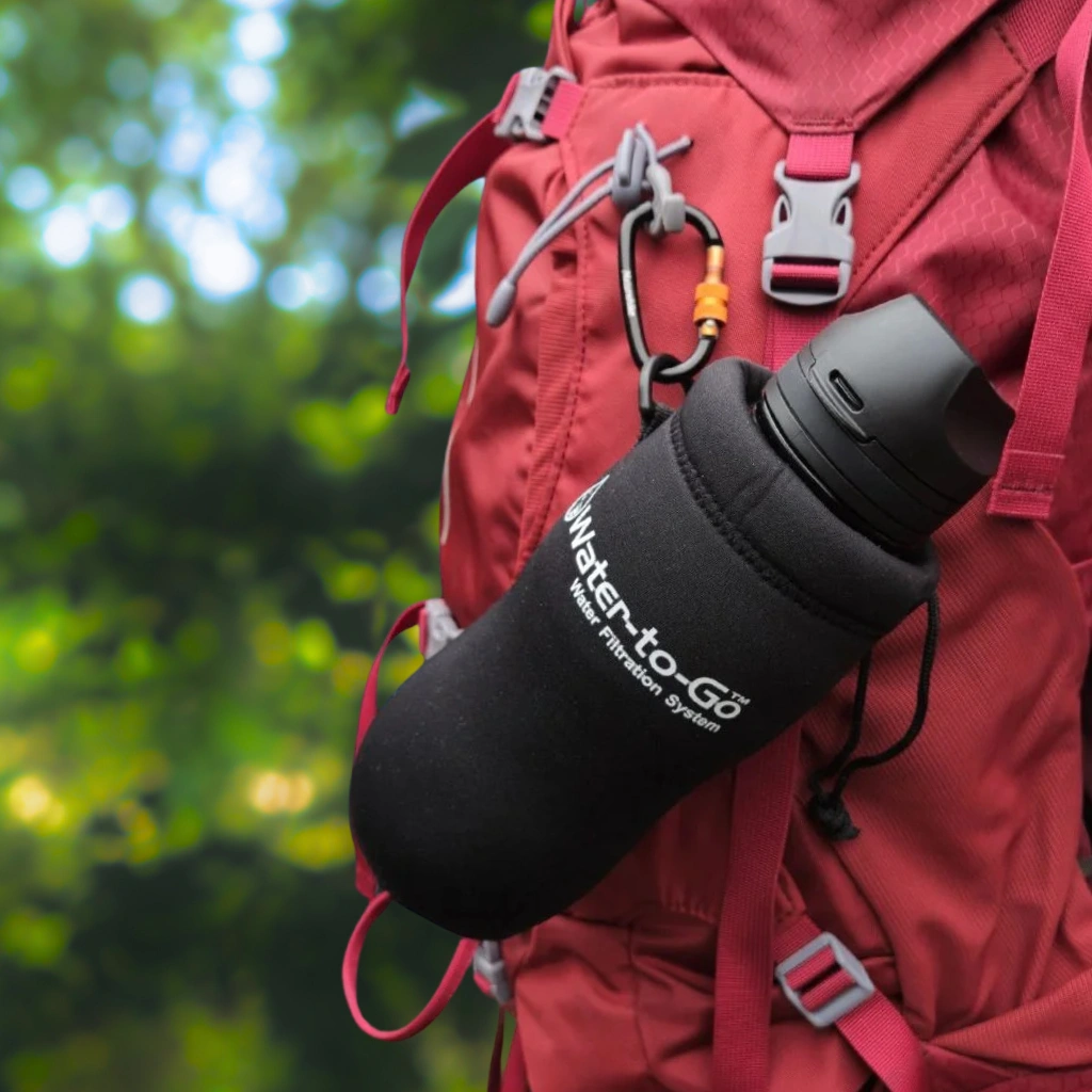 Een Water-to-Go fles met isolerende neopreen beschermhoes, bevestigd aan een backpack met een karabijnhaak. De sleeve heeft een handig oogje voor eenvoudige bevestiging.